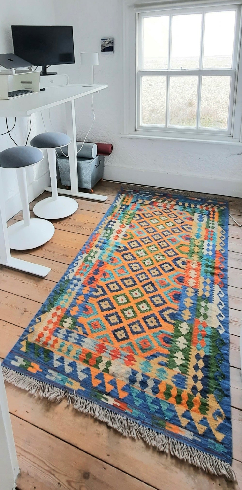 Colorful afghan kilim rug on a wooden floor with a white wall and window in the background.