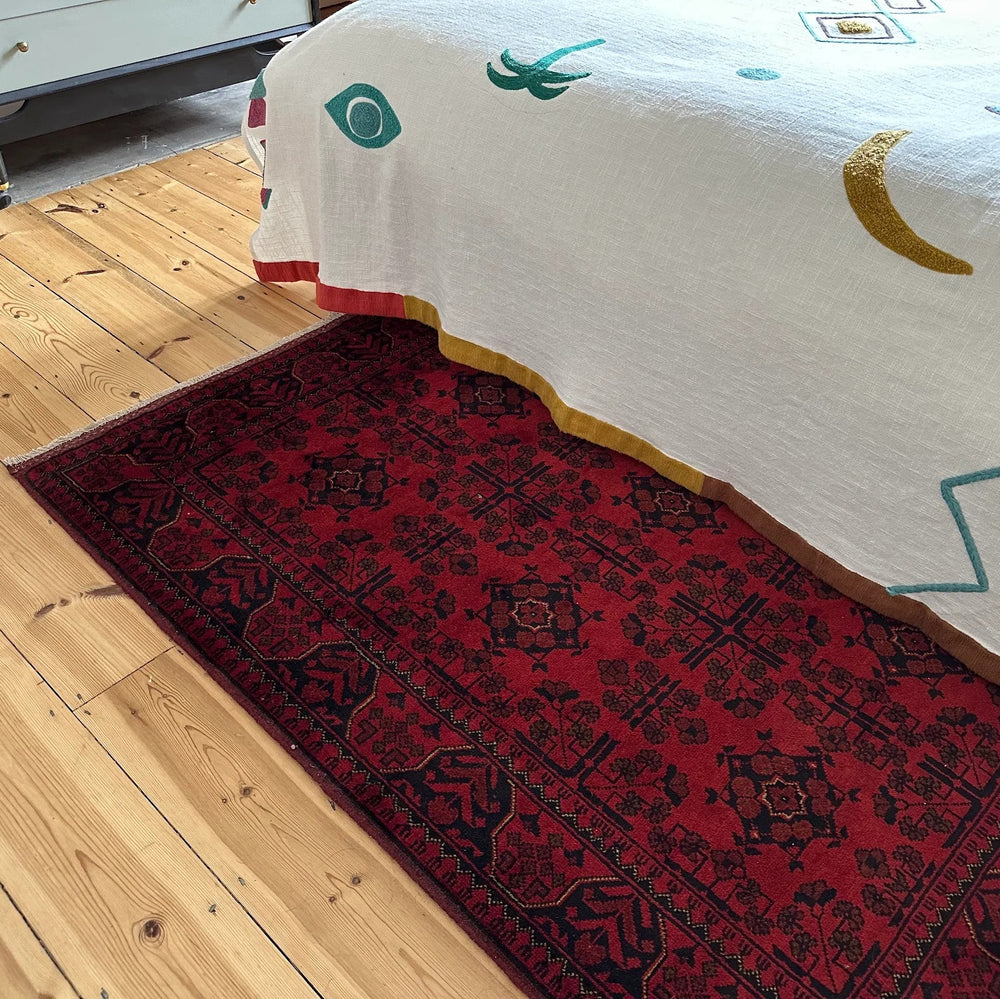Red Afghan Khal Muhammadi rug on a wooden floor next to a bed with a white comforter.