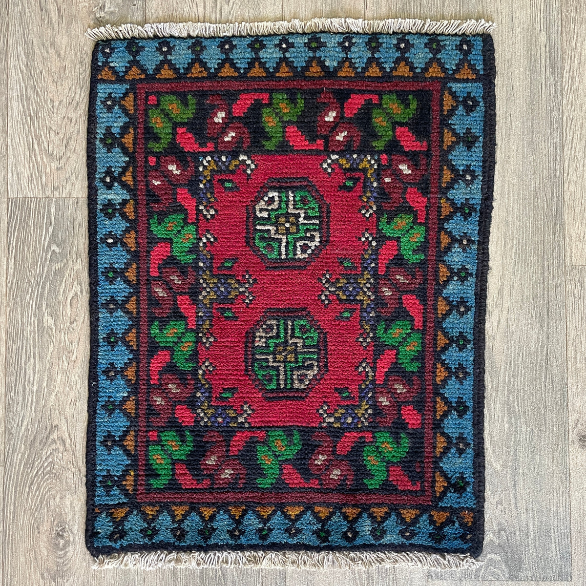 Small Afghan Aqcha handmade wool doormat featuring traditional red and navy elephant foot Mori gul patterns, styled on a light oak wood floor.