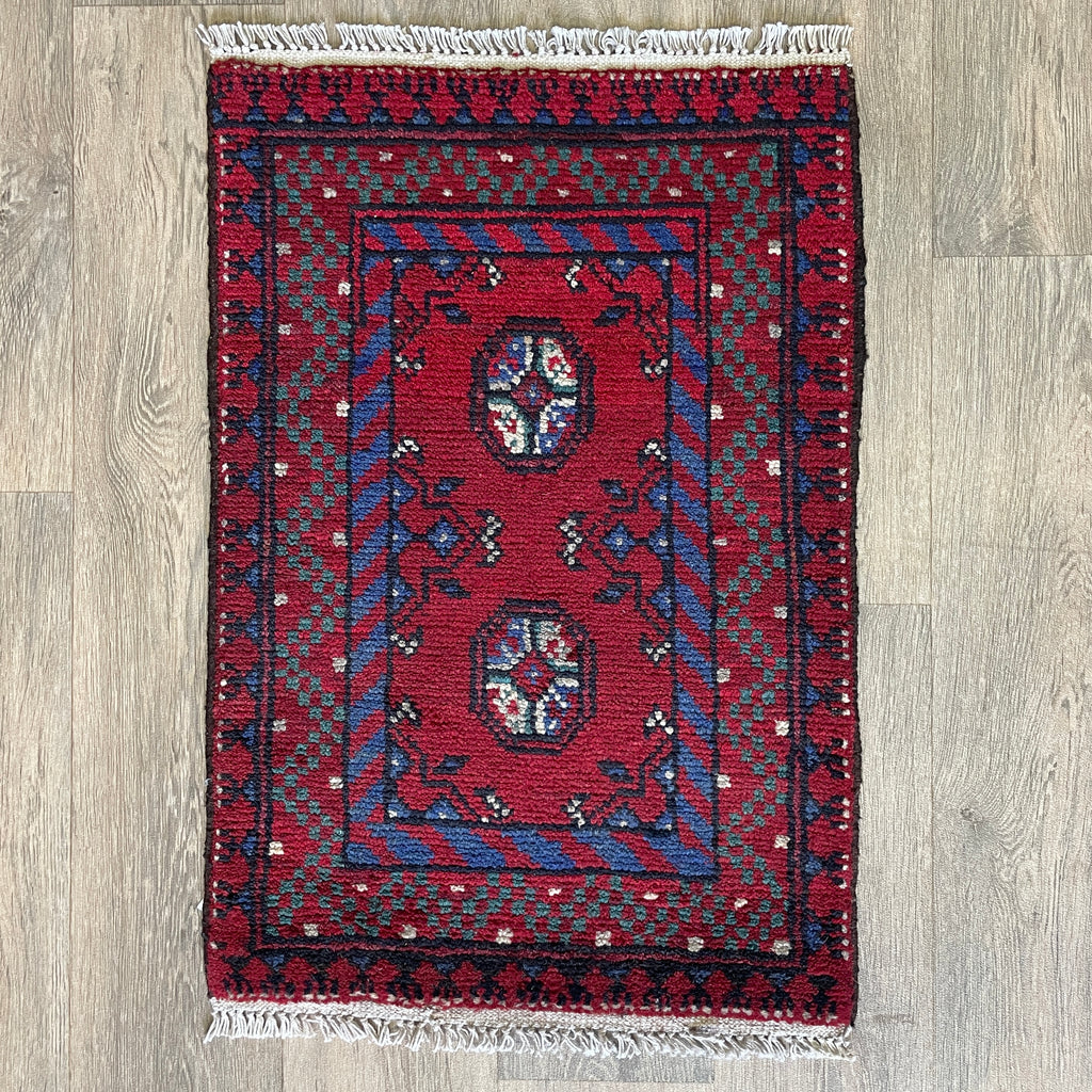 Small Afghan Aqcha handmade wool doormat featuring traditional red and navy elephant foot Mori gul patterns, styled on a light oak wood floor.