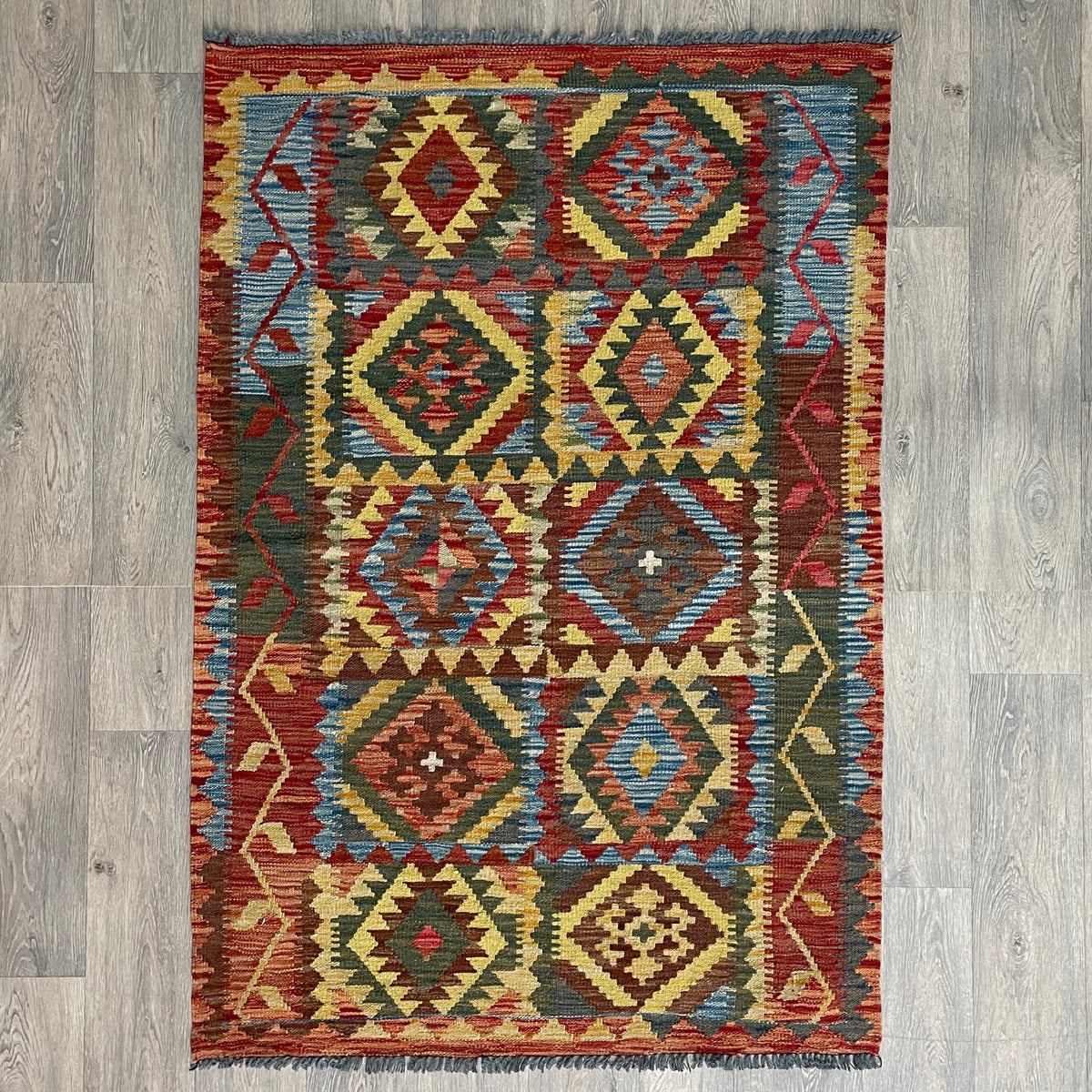 Handmade Afghan kilim flat-weave wool rug featuring bold geometric tribal patterns and braided fringes, laid flat on a light oak wood floor