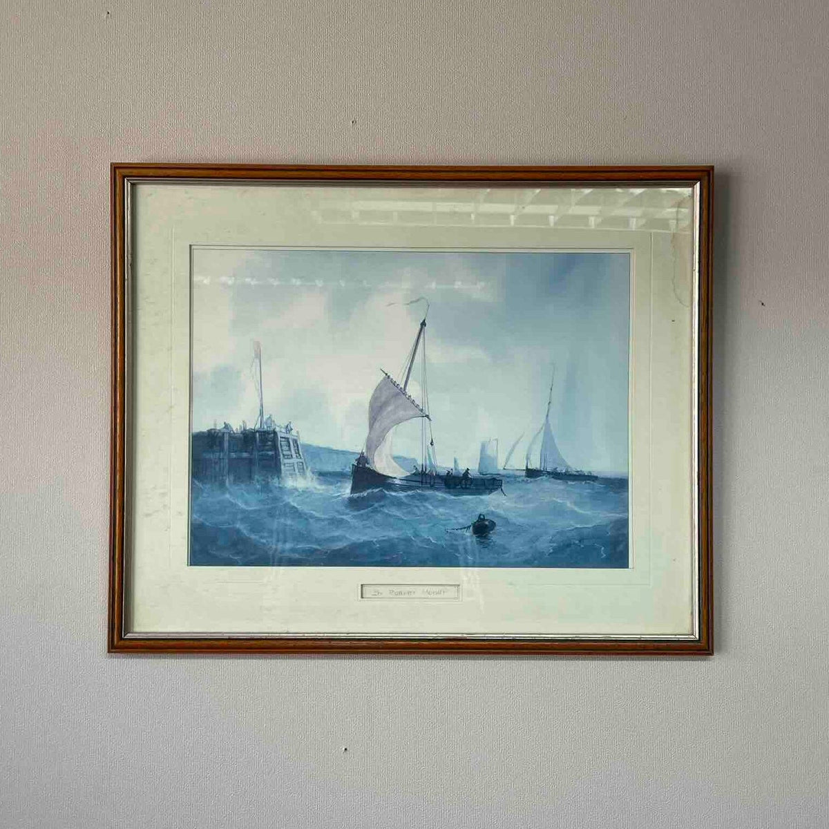 A framed artwork depicting sailboats on blue water under a cloudy sky, mounted on a light gray wall. The print has a white mat and a brown wooden frame.