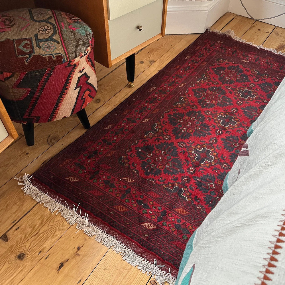 Deep red afghan khal Muhammadi rug on a wooden floor next to a bed with white bedding.